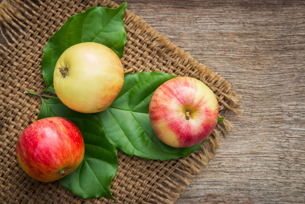 Three ripe apples on leaves over a rustic wooden background, emphasizing freshness and nutrition.